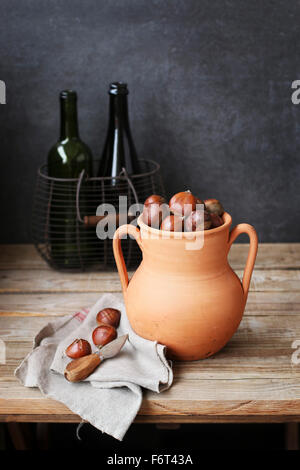 Le castagne in un rustico tostatore di argilla con due bottiglie vuote in un cesto in filo sullo sfondo Foto Stock