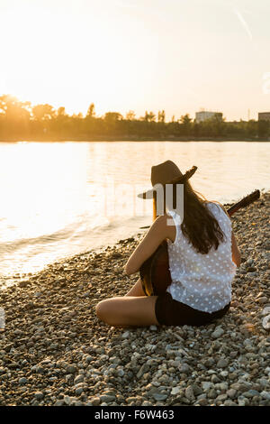 Giovane donna a suonare la chitarra in riva al fiume e al tramonto Foto Stock