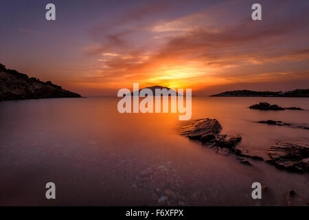 Seascape contro uno spettacolare e colorato tramonto con nuvole sognanti che passa al di sopra della baia mirroring su rocce, una lunga esposizione Foto Stock