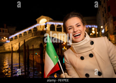 Ritratto di felice elegante giovane donna che mostra bandiera italiana mentre in piedi nella parte anteriore del ponte di Rialto in serata. Ella avente C Foto Stock