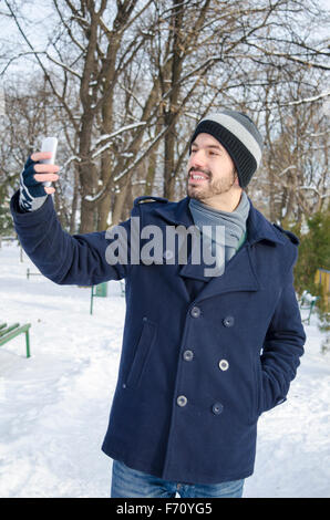 Giovane uomo barbuto prendendo un selfie in un parco su una soleggiata giornata invernale Foto Stock