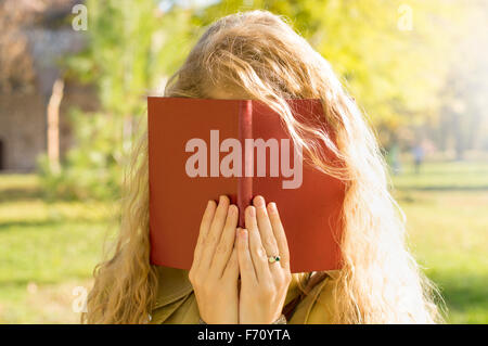 Ragazza bionda che ricopre la faccia con un libro rosso in un parco Foto Stock