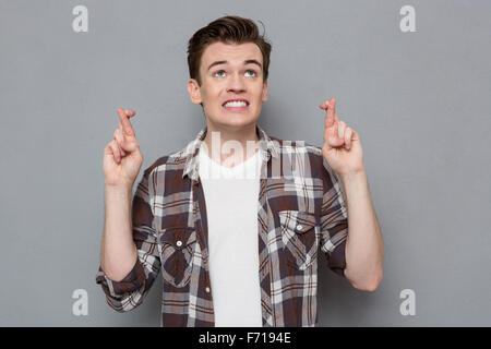 Bello concentrato giovane uomo in plaid shirt mantenendo le dita incrociate mentre in piedi contro lo sfondo grigio Foto Stock