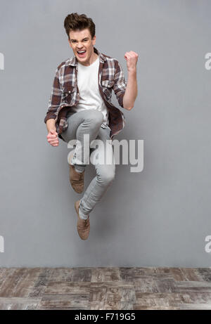 Positivo cheerfu piacere spensierato casual giovane uomo in plaid camicia e pantaloni grigi di saltare in aria e sorridente Foto Stock