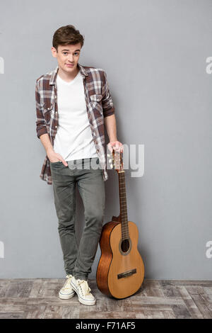 A piena lunghezza Ritratto di un giovane uomo in piedi con la chitarra su sfondo grigio Foto Stock