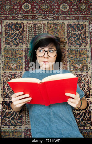 Piuttosto giovane donna sdraiata sulla moquette e la lettura di un libro rosso mentre guarda al suo fianco Foto Stock