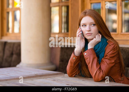 Bella pensieroso redhead giovane donna in giacca di pelle parlando al cellulare in outdoor cafe Foto Stock