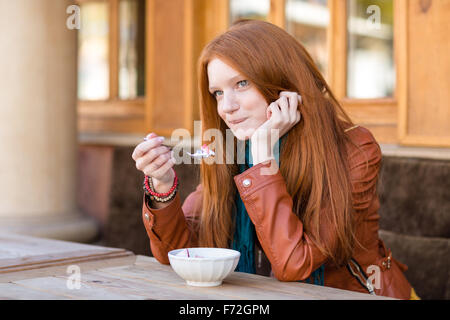 Contenuto di cute giovane donna con belle lunghi capelli rossi aventi la prima colazione nella caffetteria all'aperto e parlando al cellulare di mordere fondo li Foto Stock