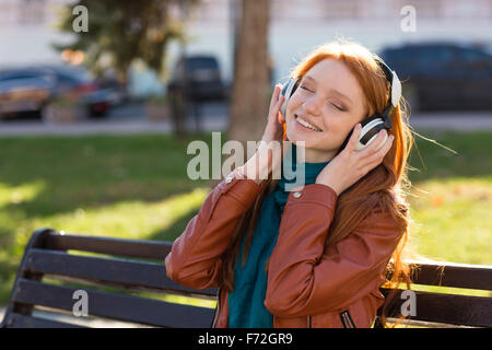 Contenuto allegro giovane donna in giacca di pelle e sciarpa seduto sulla panchina nel parco e ascolto di musica con gli occhi chiusi Foto Stock