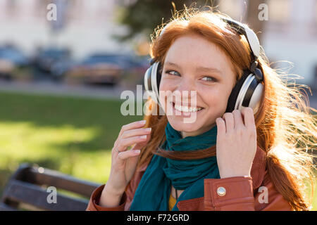 Ritratto di sorridenti redhead young lady in bright sciarpa ascoltare musica sul banco di lavoro nel parco Foto Stock