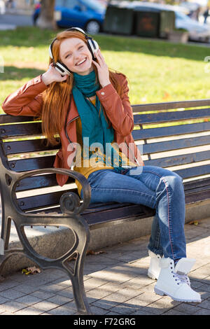 A piena lunghezza Ritratto di giovane gioiosa lieta redhead femmina in giacca di pelle, luminoso sciarpa, jeans e stivali bianco ascolto Foto Stock