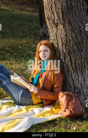 Redhead premurosa affascinante ragazza rilassante in autunno parco libro lettura sotto l'albero Foto Stock