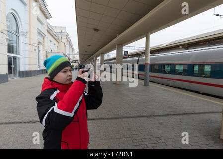 Triste ragazzo adolescente in piedi vicino al treno e fotografie sullo smartphone Foto Stock