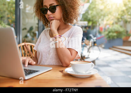 Colpo all'aperto della giovane donna africana al cafè sul marciapiede la lettura dei messaggi di posta elettronica sul suo computer portatile. Elegante ragazza seduta presso la caffetteria sur Foto Stock