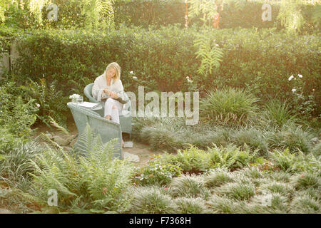 Donna bionda seduto in una sedia di vimini in un giardino, la lettura. Foto Stock