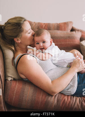 Una donna sdraiata su un divano a giocare con una bambina, baciando la sua testa. Foto Stock