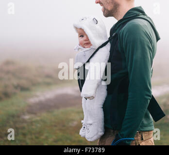 Un uomo che porta un bambino in un baby carrier sul suo petto, all'aperto in una nebbiosa giornata d'autunno Foto Stock