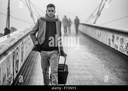 Moda uomo bello camminare su un ponte in una nebbiosa freddo giorno tirando una valigia dietro di lui come egli partono per un viaggio Foto Stock