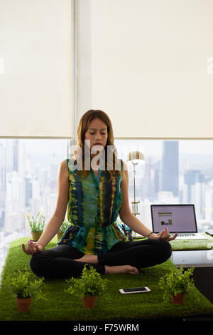 Giovane donna ispanica in ufficio, seduto sulla scrivania ricoperte di erba e piante. L'imprenditrice non lo zen e la meditazione yoga in Foto Stock