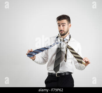 Ritratto di un confuso giovane uomo barbuto in maglia bianca la scelta giusta cravatta oltre il grigio di sfondo per studio Foto Stock