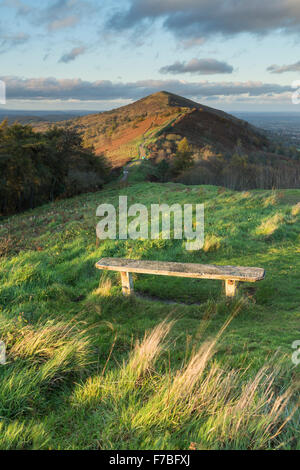 Una panca in legno sulla collina Jubillee accesa dal bel tramonto che è anche illuminazione di perseveranza Hill e il Worcestershire Beacon Foto Stock