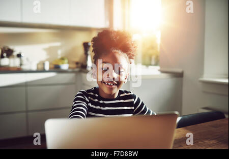 Carino giovane ragazza nera la riproduzione su un computer portatile sorridente felicemente come Lei naviga in internet mentre vi rilassate in un tavolo a casa, Foto Stock