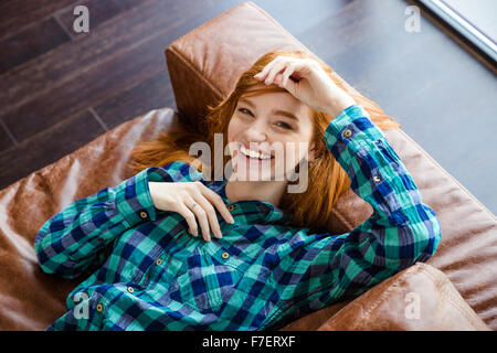 Bella naturale gioiosa giovane donna in maglia a scacchi posa su divani in pelle marrone e ridere Foto Stock