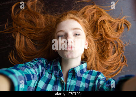 Vista dall'alto di attraenti giocosa donna giovane con bellissimi capelli rossi in camicia a scacchi facendo selfie e di inviare aria kiss giacente o Foto Stock