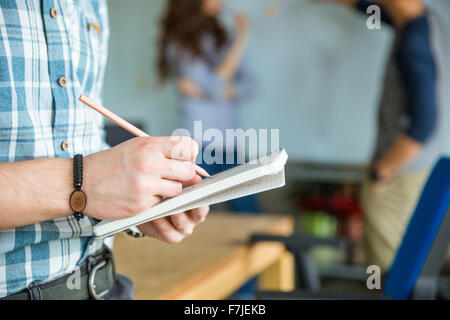 Le mani di uomo in camicia a scacchi la scrittura in notebook dalla matita mentre i colleghi a discutere di un nuovo progetto Foto Stock