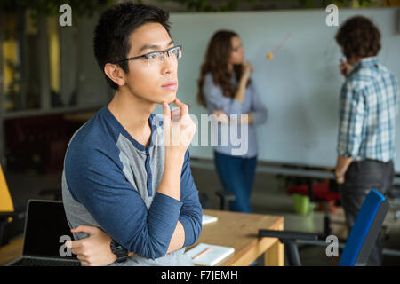 Malinconici concentrata bel giovane uomo asiatico in bicchieri di pensare e di lavorare con i colleghi Foto Stock