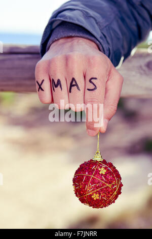 Primo piano di un giovane uomo caucasico con la parola xmas scritto in le nocche della mano tenendo un rosso palla di Natale all'aperto Foto Stock
