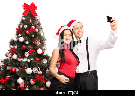 Giovane uomo e donna con cappelli di Babbo Natale prendendo un selfie nella parte anteriore di un albero di Natale isolato su sfondo bianco Foto Stock