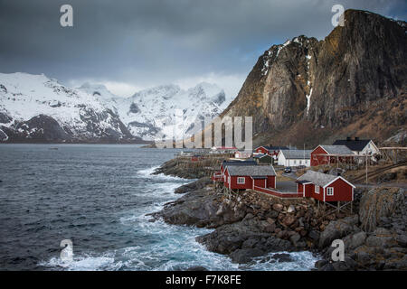 Montagne coperte di neve dietro erte villaggio di pescatori sulla baia, Hamnoya, Isole Lofoten in Norvegia Foto Stock