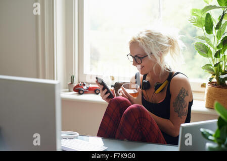 Giovane Donna con cuffie e tatuaggio texting in home office Foto Stock