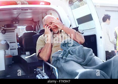 Uomo tormentato sulla barella parlando al cellulare dietro ambulanza Foto Stock