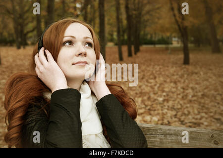 Redhead girl ascoltare musica su cuffie in city park, caduta stagione Foto Stock