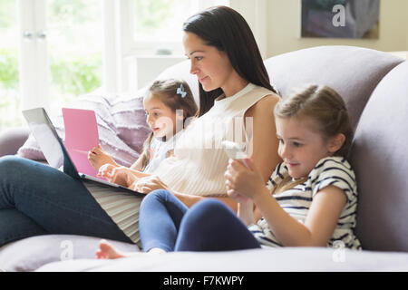 La madre e le figlie con il computer portatile, telefono cellulare e tavoletta digitale sul divano Foto Stock