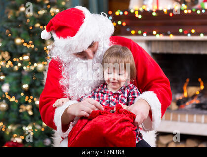 Babbo Natale dando un presente dal sacco al bambino ragazzo vicino al camino e albero di Natale a casa. Foto Stock