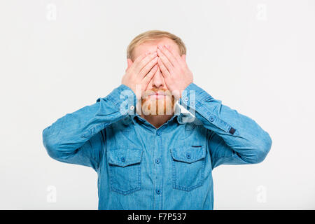 Ritratto di giovane bella bionda maschio barbuto in jeans shirt con gli occhi coperti da mani su sfondo bianco Foto Stock