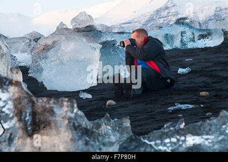 Fotografo con blocchi di ghiaccio sulla spiaggia presso Jokulsarlon glaciale, spiaggia spiaggia di Diamante, con montagne coperte di neve in lontananza, in Islanda in febbraio Foto Stock
