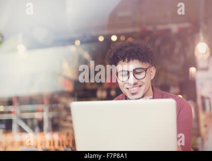 Uomo sorridente con ricci capelli neri utilizzando computer portatile presso il cafe finestra Foto Stock