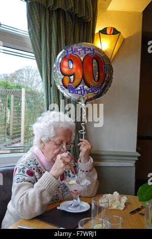 Una donna anziana che celebra il suo novantesimo compleanno di mangiare un gelato Gelato e tenendo un palloncino con felice il novantesimo compleanno su di esso, England, Regno Unito Foto Stock