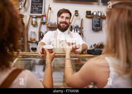 Gentile giovane uomo con la barba in piedi dietro il cafe contatore e che serve caffè per due donne. Giovane uomo che lavora nel ristorante Foto Stock