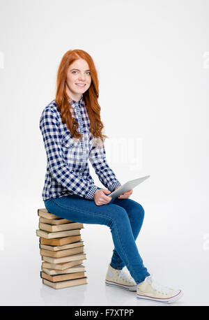 Allegro positivo readhead ricci lady in plaid shirt e jeans seduti sulla pila di libri e utilizzo di tablet su sfondo bianco Foto Stock