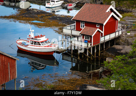 Rosso mattone cabina in legno su palafitte e piccola barca da pesca vicino al porto di Moskenes in western Isole Lofoten in Norvegia Foto Stock
