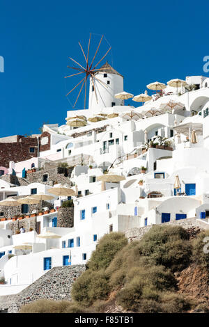 Santorini, la cittadina di Oia. È possibile visualizzare fino a imbiancato cubiform edifici costruiti in scogliera-faccia, con tipico mulino a vento sull orizzonte contro il cielo blu e chiaro. Foto Stock