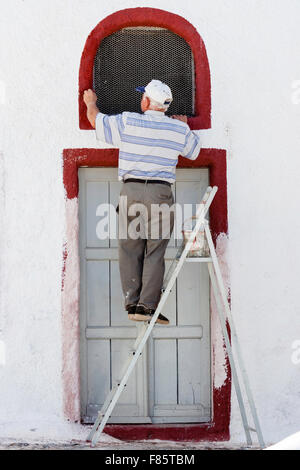 Santorini. Greco Senior uomo in piedi sul gradino-scala, all'esterno, la verniciatura del telaio della finestra cremisi scuro al di sopra di porta nella parete dipinta di bianco. Fai da te. Foto Stock