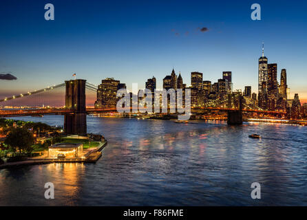 Il Ponte di Brooklyn e illuminato la parte inferiore di Manhattan al crepuscolo. Il quartiere finanziario di grattacieli riflettono in East River, New York. Foto Stock