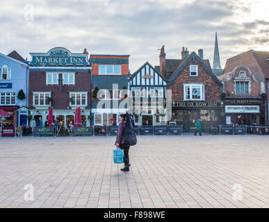 Uomo che cammina con una chitarra sulla schiena in tutta la piazza del mercato, Salisbury, Wiltshire, Regno Unito. La guglia della Cattedrale di Salisbury, il più alto in Inghilterra, è visibile al di sopra di negozi e ristoranti. Foto Stock