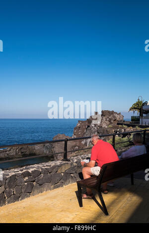 I turisti seduta su una panchina lungo la passeggiata pedonale in Puerto Santiago, Tenerife, Isole Canarie, Spagna. Foto Stock